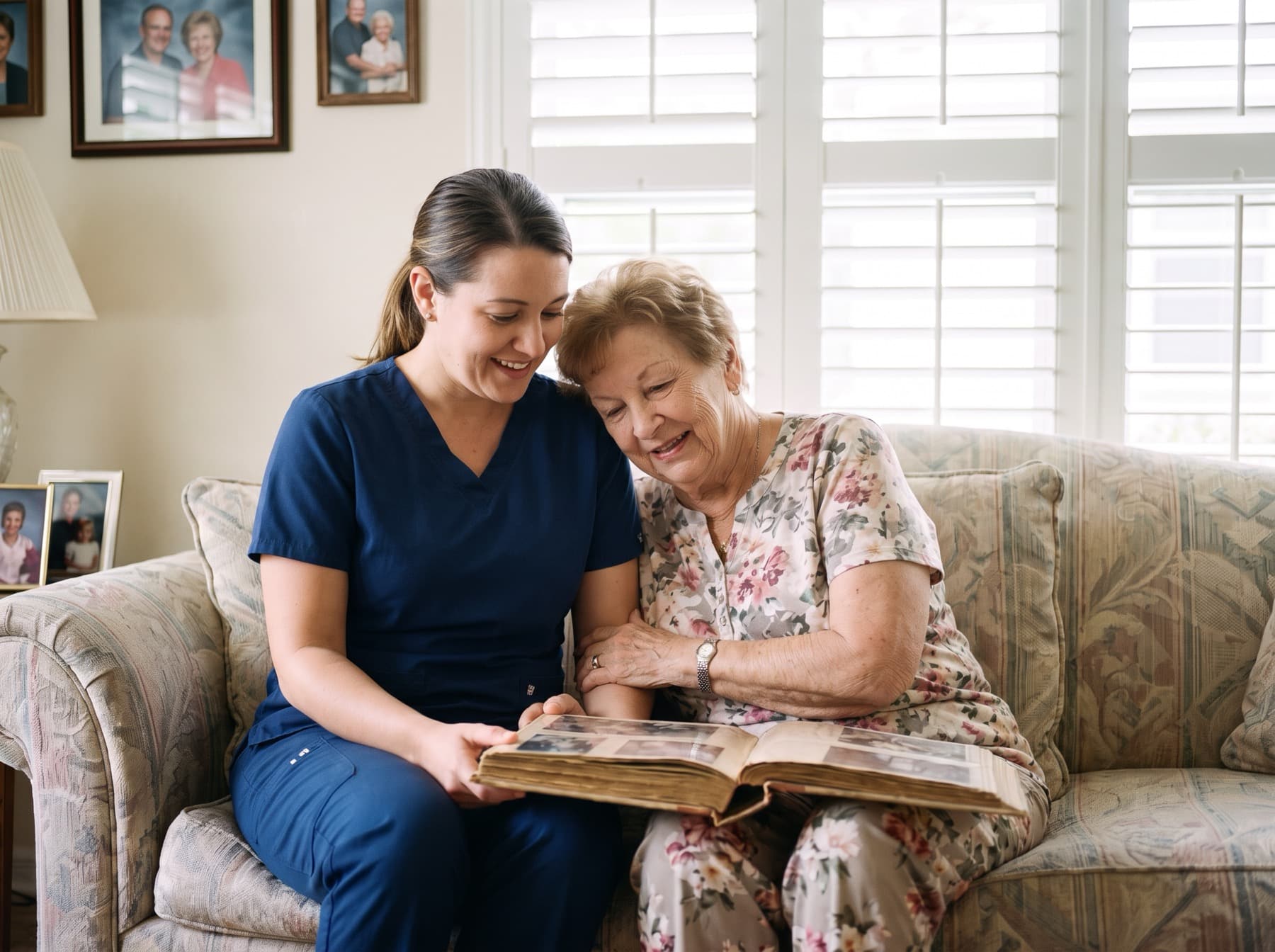 A caregiver in navy scrubs looking through an old photo album with an older woman leaning in beside her on the sofa.